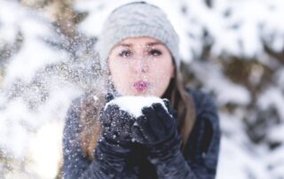 A woman is blowing snow towards the camera while being in the snow, surrounded by trees.