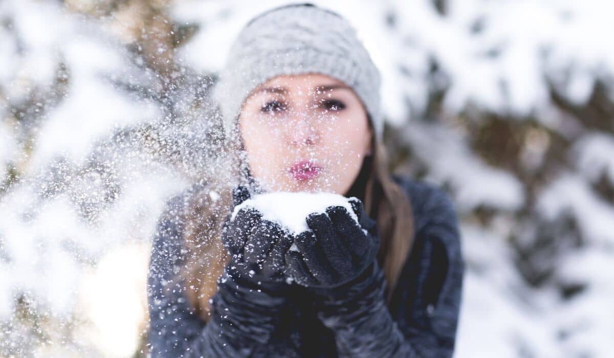 A woman is blowing snow towards the camera while being in the snow, surrounded by trees.